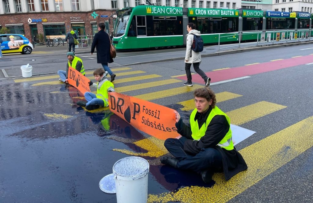 Am 18. April störten Unterstützer/innen von DROP Fossil Subsidies den Morgenverkehr in Basel in der Nähe des Bahnhofs SBB. Sie schütteten eine ölähnliche Flüssigkeit auf die Fahrbahn, setzten sich hin und breiteten orange Banner mit der Aufschrift «DROP Fossil Subsidies» aus. Angesichts der Klimakrise fordert die Bewegung den Bundesrat auf, einen Plan zur sofortigen Beendigung aller fossilen Subventionen aufzustellen. «Indem weiterhin Milliarden von Schweizer Franken in die Subventionierung fossiler Brennstoffe gesteckt werden, versagt die Regierung in ihrer Fürsorgepflicht gegenüber den Bürgern/innen und verstösst, wie der EGMR bestätigt, gegen deren Menschenrechte», so DROP Fossil Subsidies in einer Pressemitteilung. | ©DROP Fossil Subsidies  